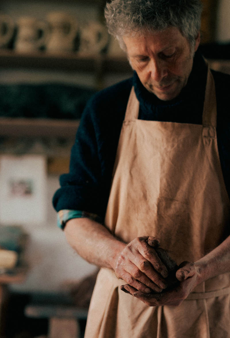James with lowered head ball of clay in hands, jugs in background